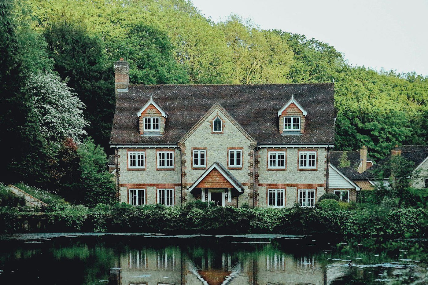 A stone and brick house with a tiled roof, reflected in still water and backed by trees
